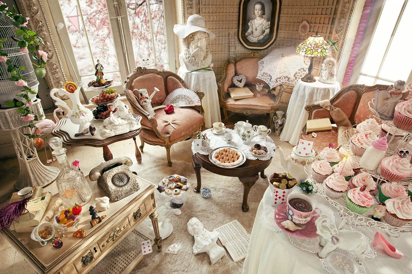 Sala decorada con muebles antiguos y objetos decorativos, incluyendo tazas de té, pasteles, cupcakes rosados, porcelanas, una máquina de escribir, una figura de perro de cerámica, y decoraciones vintage en colores pastel y rosa.